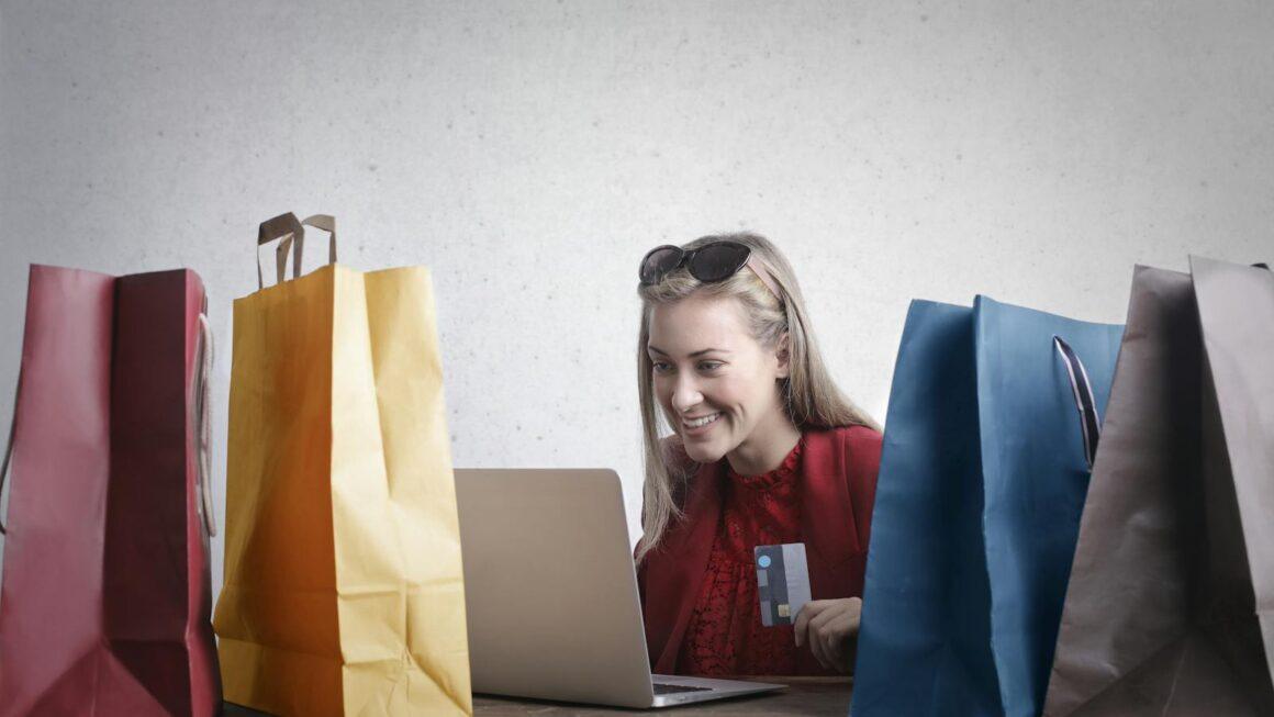 A joyful woman shopping online using a laptop with colorful shopping bags surrounding her.