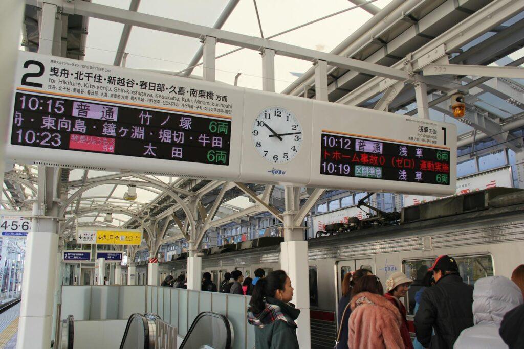 Passengers waiting at a Japanese train platform with a clock and timetable displays.